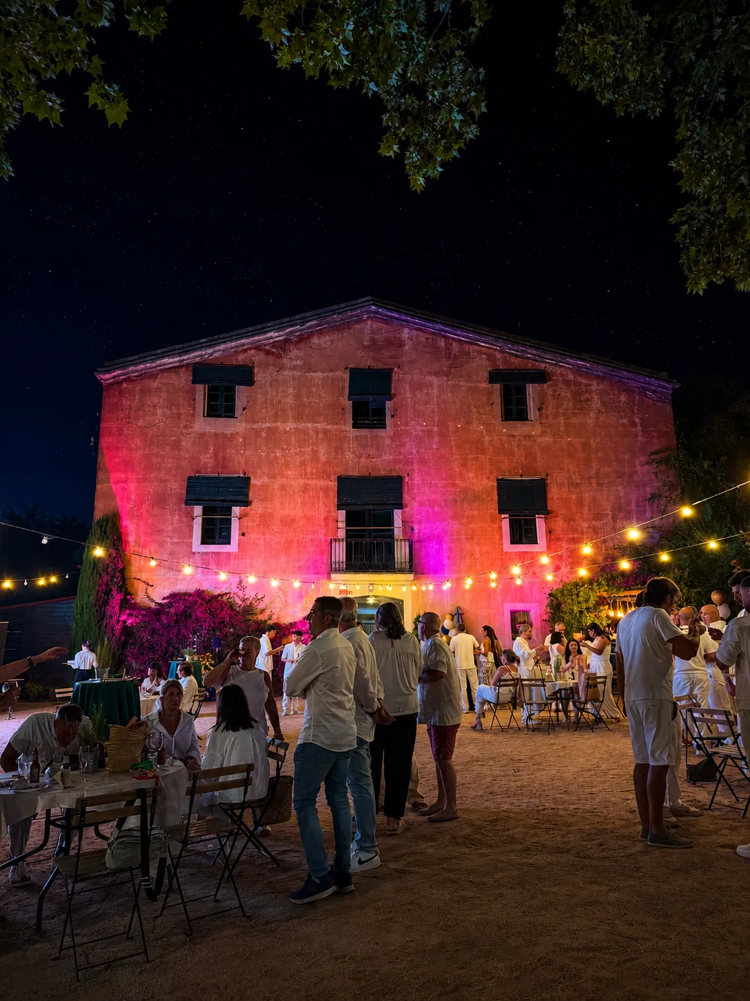 Masía de Alella iluminada en tonos cálidos de noche durante el evento corporativo, con guirnaldas e invitados vestidos de blanco